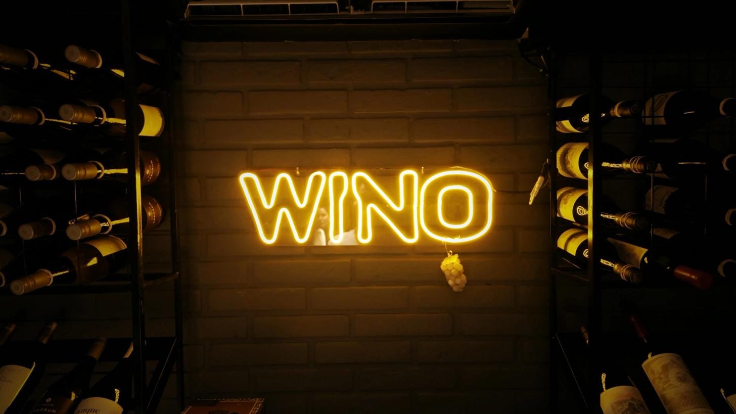 WINO's neon sign glowing on a white brick wall in the wine cellar, framed by racks of bottles on both sides. WINO's neon sign glowing on a white brick wall in the wine cellar, framed by racks of bottles on both sides.