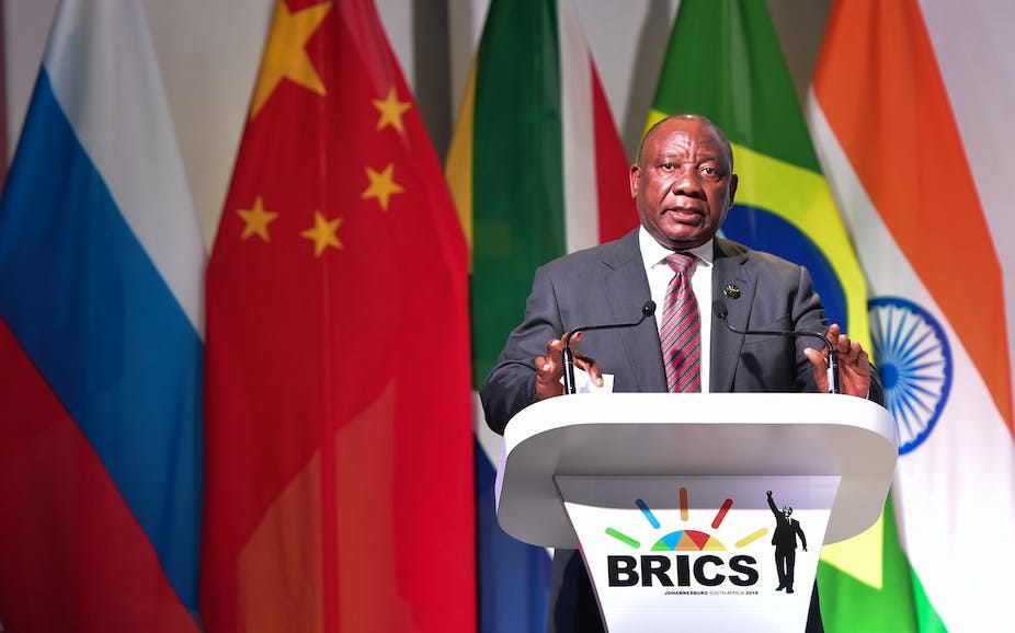 A man wearing a suit and tie speaks at a podium bearing the BRICS logo. A man wearing a suit and tie speaks at a podium bearing the BRICS logo.