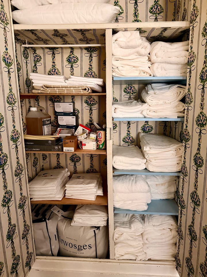 A woman walking through a furnished coach house sitting room; an open cupboard of folded white linens and towels; a woman in a straw hat clipping hydrangeas; a blue-and-white vase of pink and purple hydrangeas.