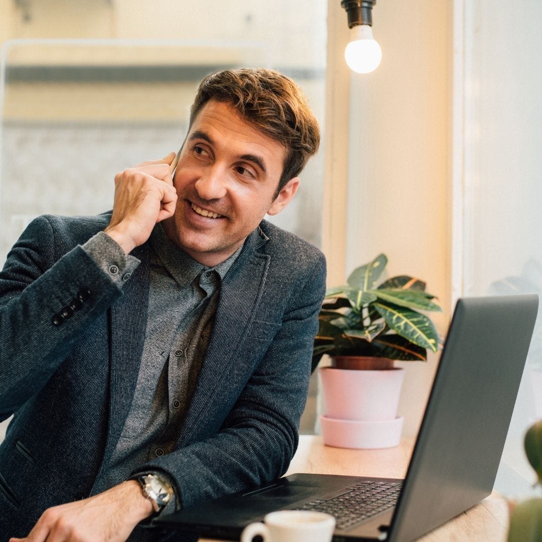 Man leaning on desk with laptop and coffee, one hand holding a phone to his ear as he smiles.