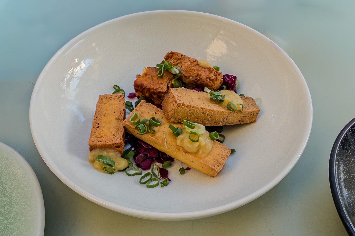 Weisse Schüssel mit mehreren Stücken Räuchertofu auf fermentiertem Rotkohl. Dazu kommen helle Creme-Tupfen und fein geschnittene Frühlingszwiebeln, die dem Gericht auf dem hellen Tisch grüne und violette Akzente geben. Weisse Schüssel mit mehreren Stücken Räuchertofu auf fermentiertem Rotkohl. Dazu kommen helle Creme-Tupfen und fein geschnittene Frühlingszwiebeln, die dem Gericht auf dem hellen Tisch grüne und violette Akzente geben.