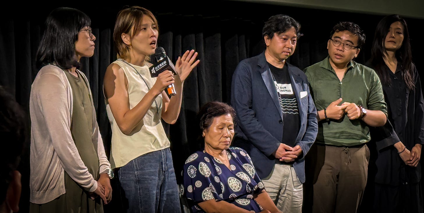 A woman in a beige tanktop and jeans speaks into a microphone. She is surrounded by multiple people, including a seated elderly woman with downcast eyes