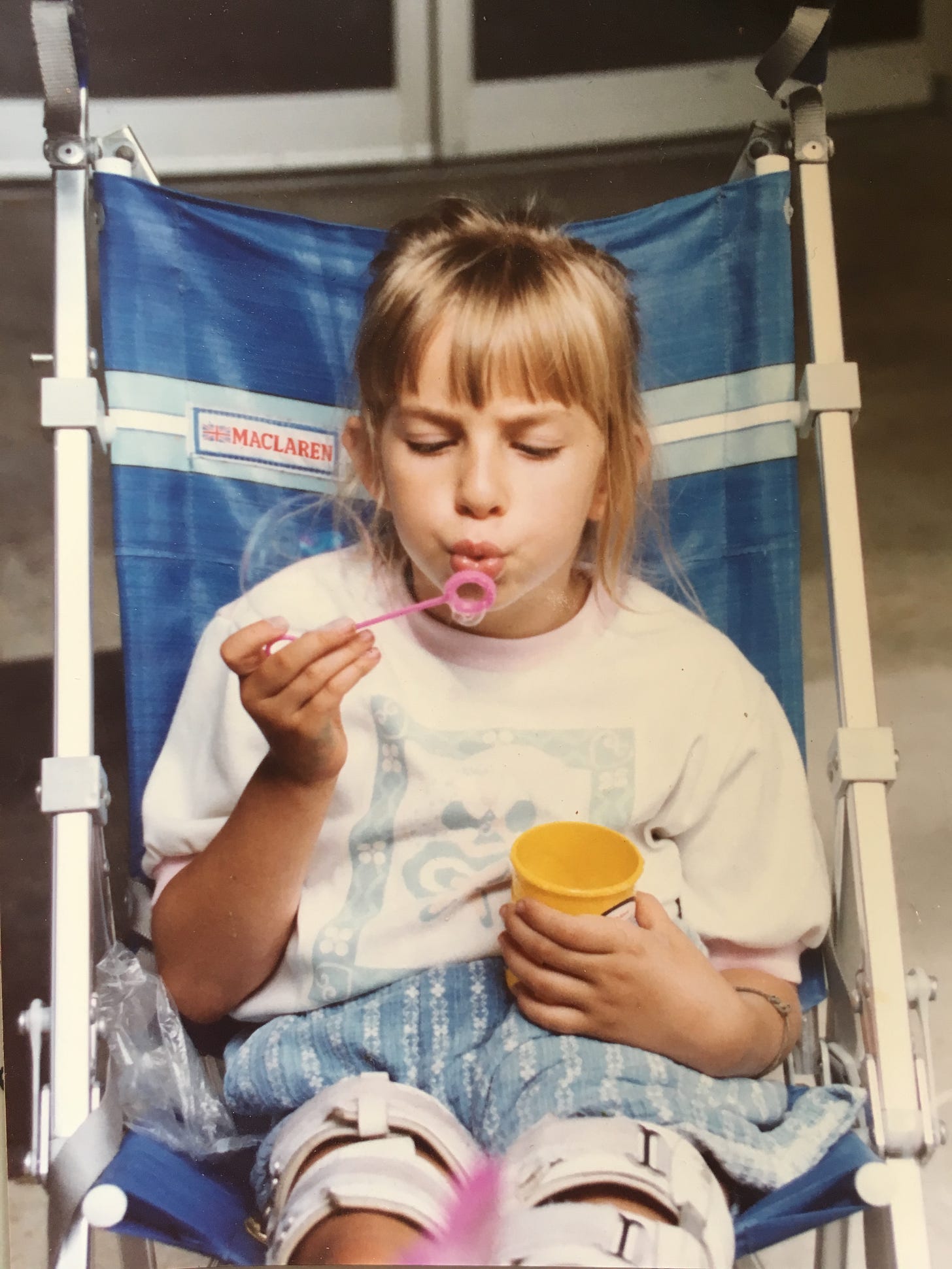 A young Rebekah sits in a blue stroller, leg braces up to her thighs. She furrows her brow as she concentrates on blowing soap bubbles through a pink wand. A young Rebekah sits in a blue stroller, leg braces up to her thighs. She furrows her brow as she concentrates on blowing soap bubbles through a pink wand.