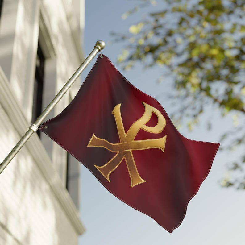 May include: A burgundy flag with a gold Chi Rho symbol, a monogram of the first two letters of the Greek word for Christ. The flag is waving on a silver pole, mounted on a building with a white facade. The background includes a blue sky and green foliage.