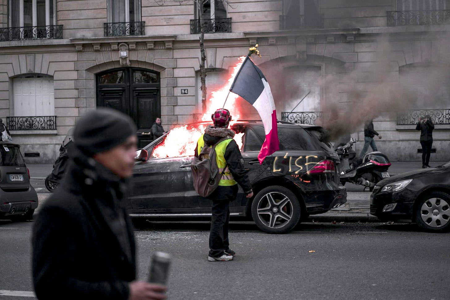 The image shows a protest scene in Paris with a Yellow Vest demonstrator holding a French flag next to a luxury car that's on fire. The burning vehicle has "LISE" spray-painted on its side. A passerby in dark clothing walks in the foreground, while Parisian buildings form the backdrop. The scene captures civil unrest during Yellow Vest movement protests in France.