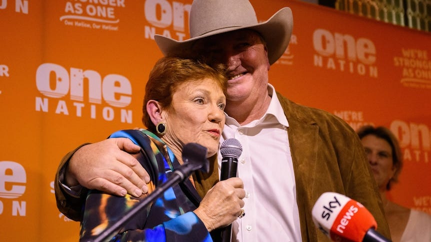 A man in an Akubara wraps his arms around a woman with a microphone.
