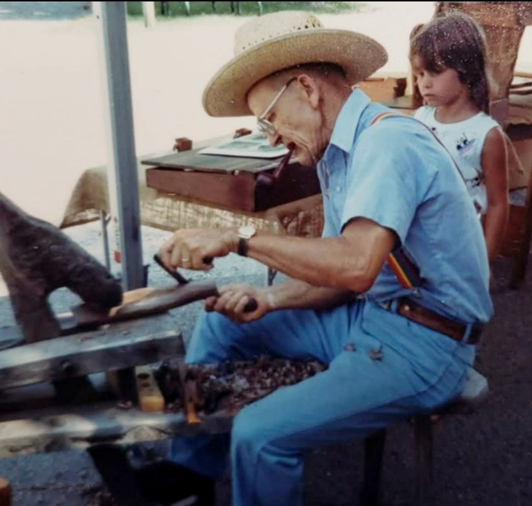 An elderly man in a straw hat and blue work clothes uses a drawknife on a shaving horse, shaping a wood blank, while a young girl watches from behind
