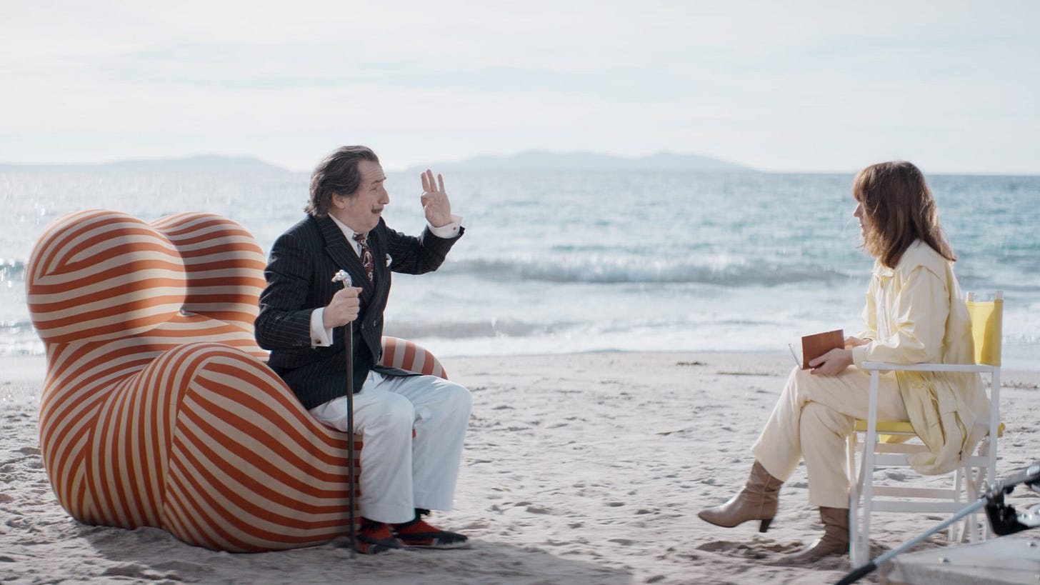 Two people sitting on a beach facing each other, one in a striped chair, the other in a folding chair, with the ocean in the background.