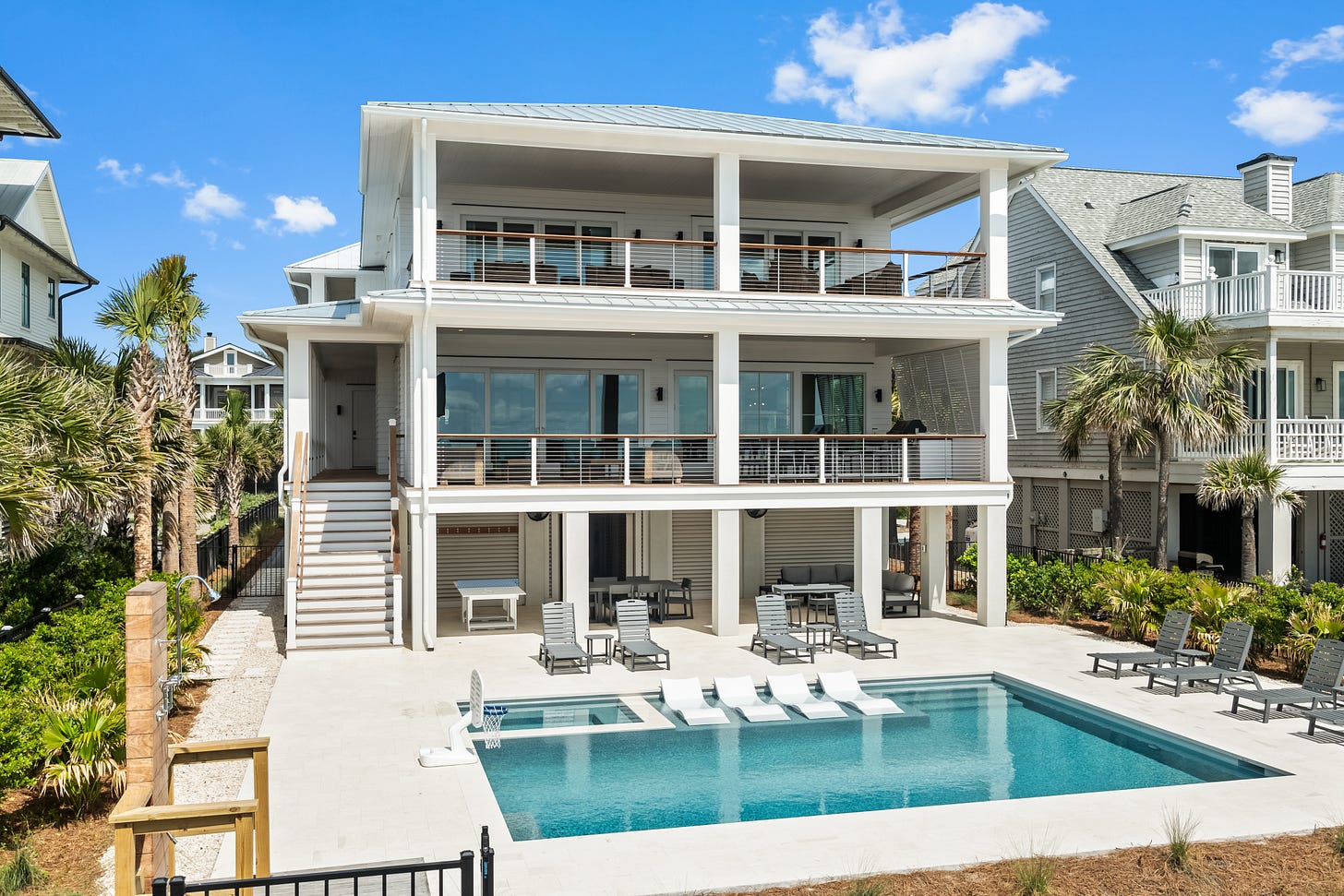 Rear exterior of a three-story white coastal house with a metal roof, covered porches on two levels, and a rectangular pool surrounded by lounge chairs and low landscaping. Rear exterior of a three-story white coastal house with a metal roof, covered porches on two levels, and a rectangular pool surrounded by lounge chairs and low landscaping.