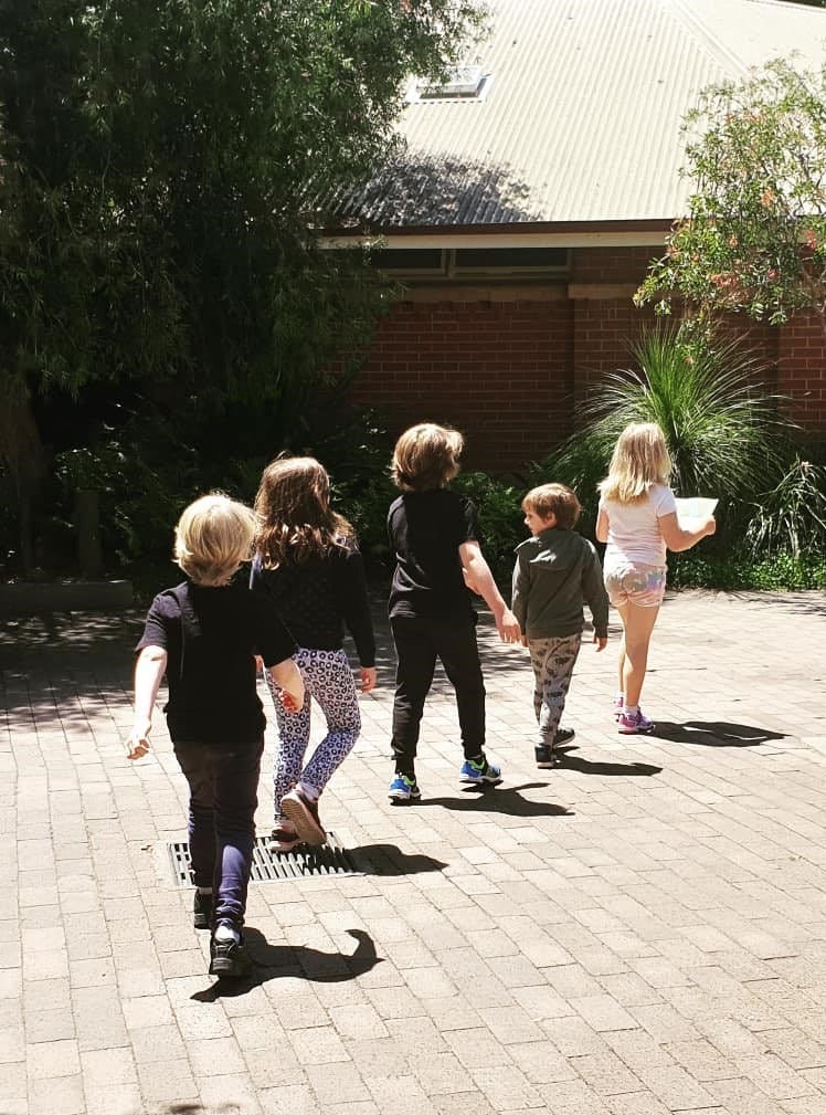 five children aged between 6 and 11 walking in a line with the leader holding a map, along a paved path between shrubs towards a well-shaded building