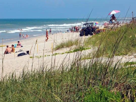 People relax and walk along the shoreline at Playalinda Beach on the Canaveral National Seashore, with lifeguard stands, beach umbrellas, and dune grass visible in the foreground. People relax and walk along the shoreline at Playalinda Beach on the Canaveral National Seashore, with lifeguard stands, beach umbrellas, and dune grass visible in the foreground.