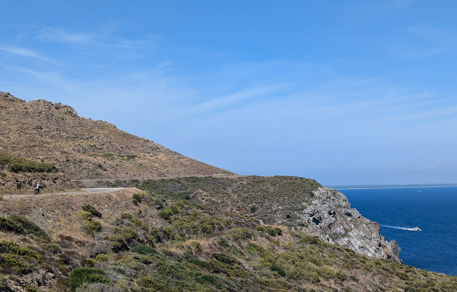 Vegetated cliffs overlooking a deep blue sea and blue skies, a cyclist on the road which curves around the cliffs
