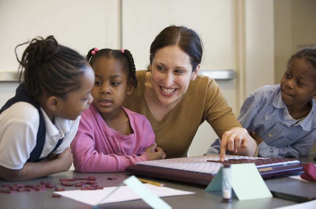 A teacher smiles as she plays Scrabble with three little girls, pointing out something on the board to one of the kids. They all seem engaged and excited to learn. A teacher smiles as she plays Scrabble with three little girls, pointing out something on the board to one of the kids. They all seem engaged and excited to learn.