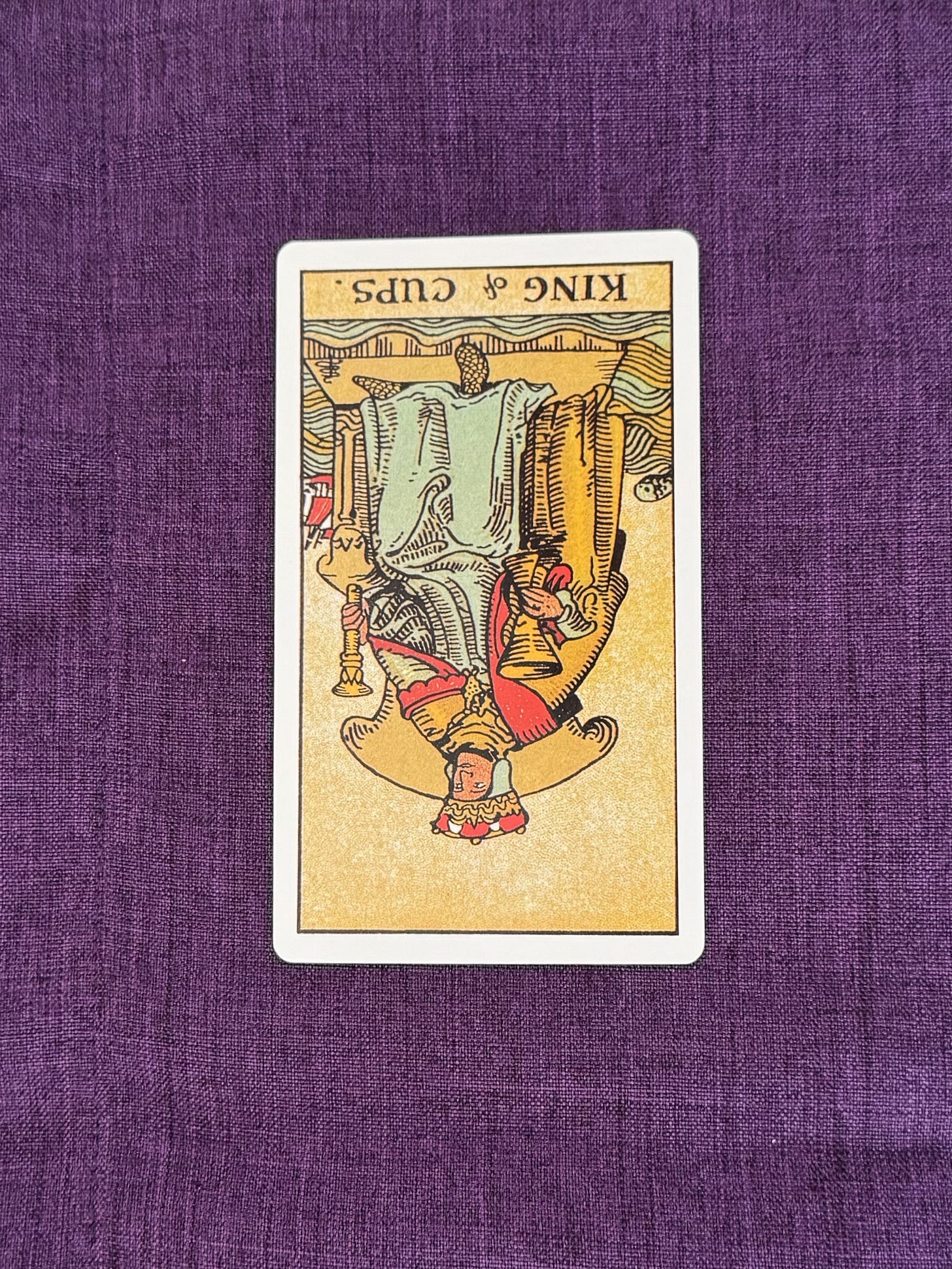 Tarot Card is placed upsidedown on a purple linen cloth. The image depicts a white haired, white man, sitting on a throne, wearing a crown, holding a cup in his right hand and a septre in his left.