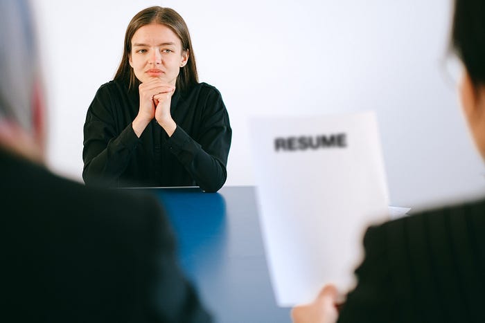 A woman with her elbows on the table and hands under her head, looking pensively at another person holding a piece of paper that says Resume A woman with her elbows on the table and hands under her head, looking pensively at another person holding a piece of paper that says Resume