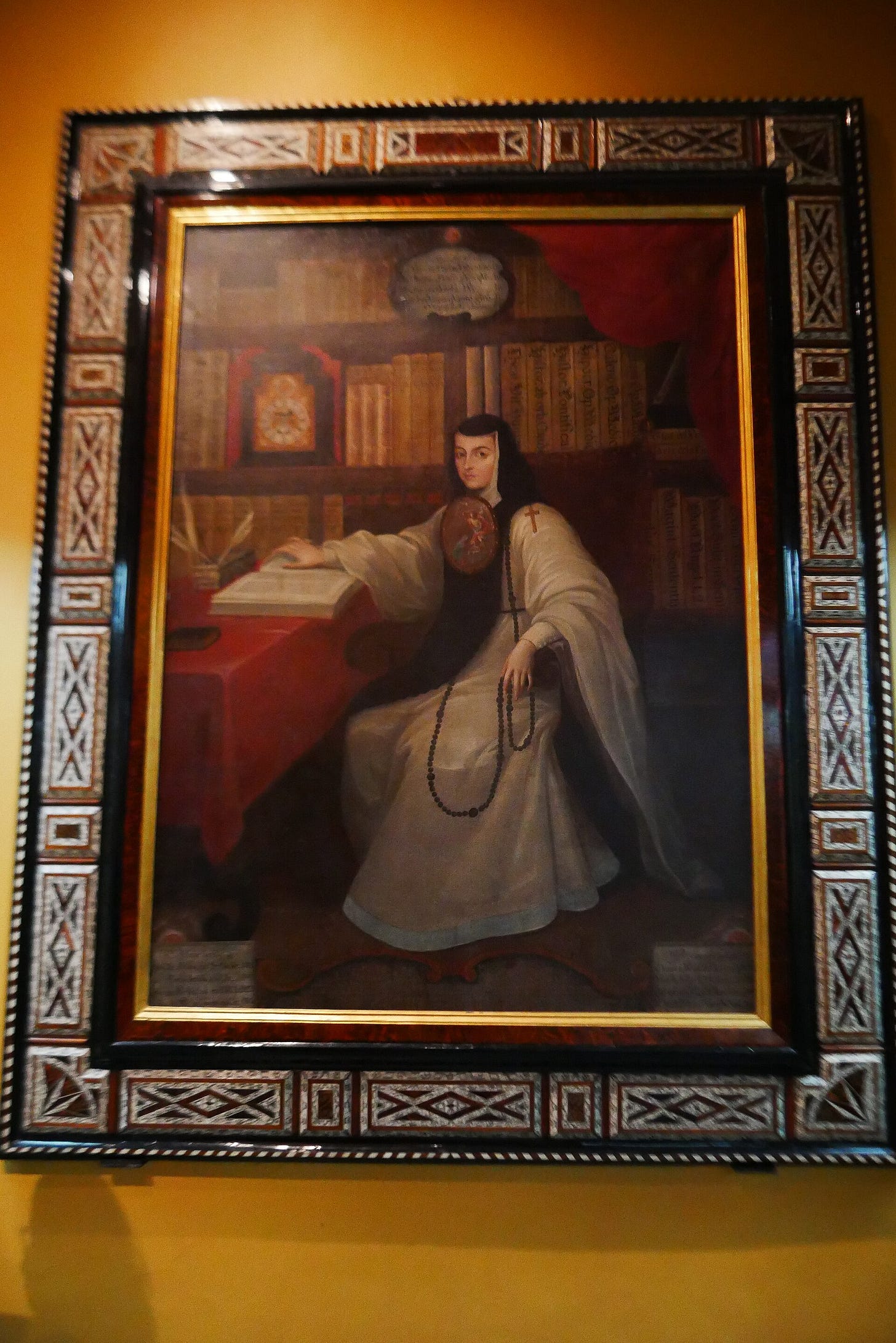 A nun in a white habit with a large painted badge on her chest sits at a desk, turning the page of a book, in front of shelves of leather volumes and a red curtain.