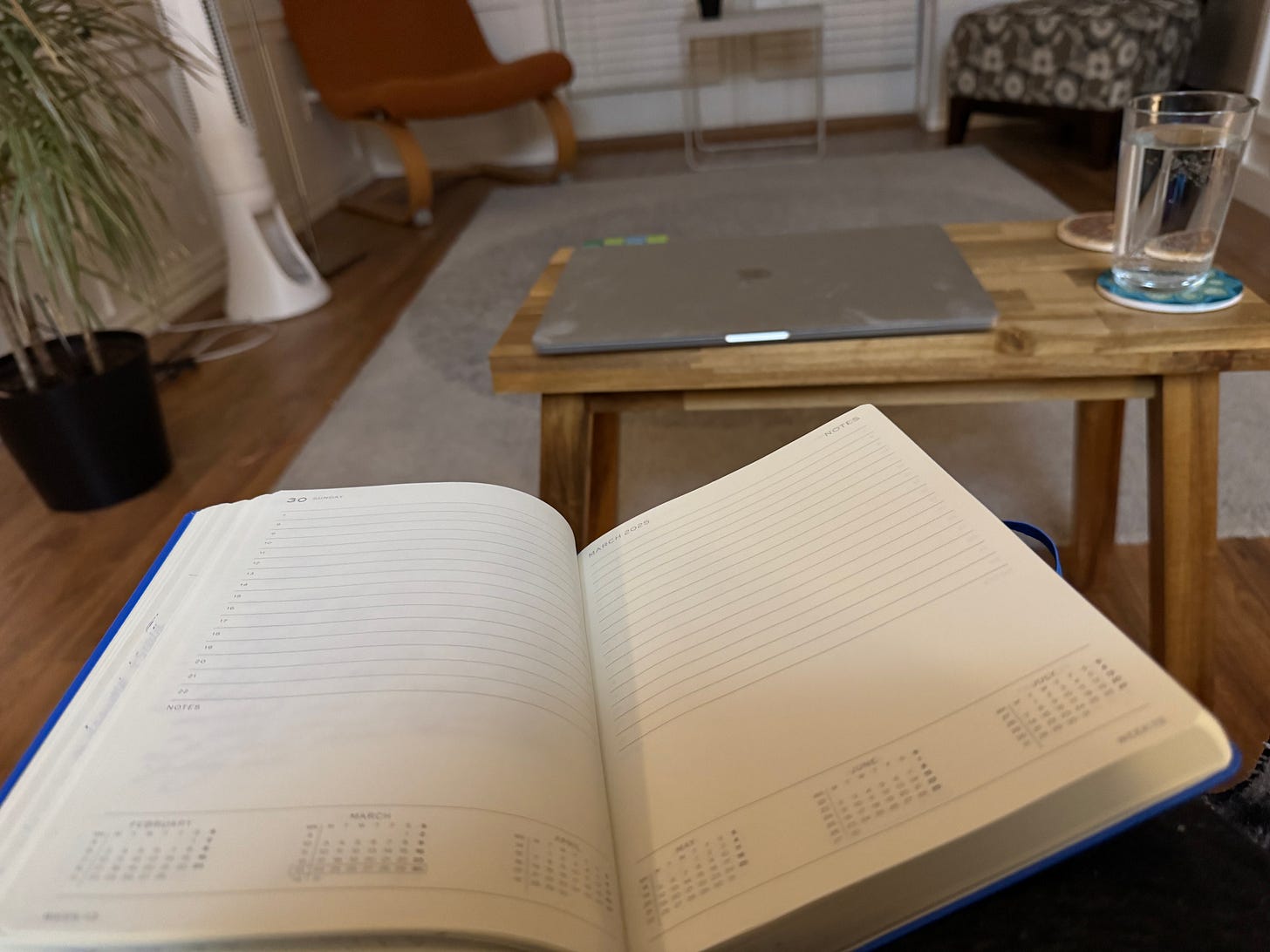 Day timer open on my lap in a living room beside a small wooden coffee table where a laptop sits beside a glass of water.