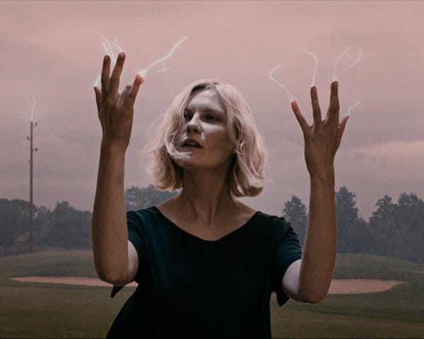 A blonde woman in a dark outfit holds her spread fingers up in the air against a pink sky.