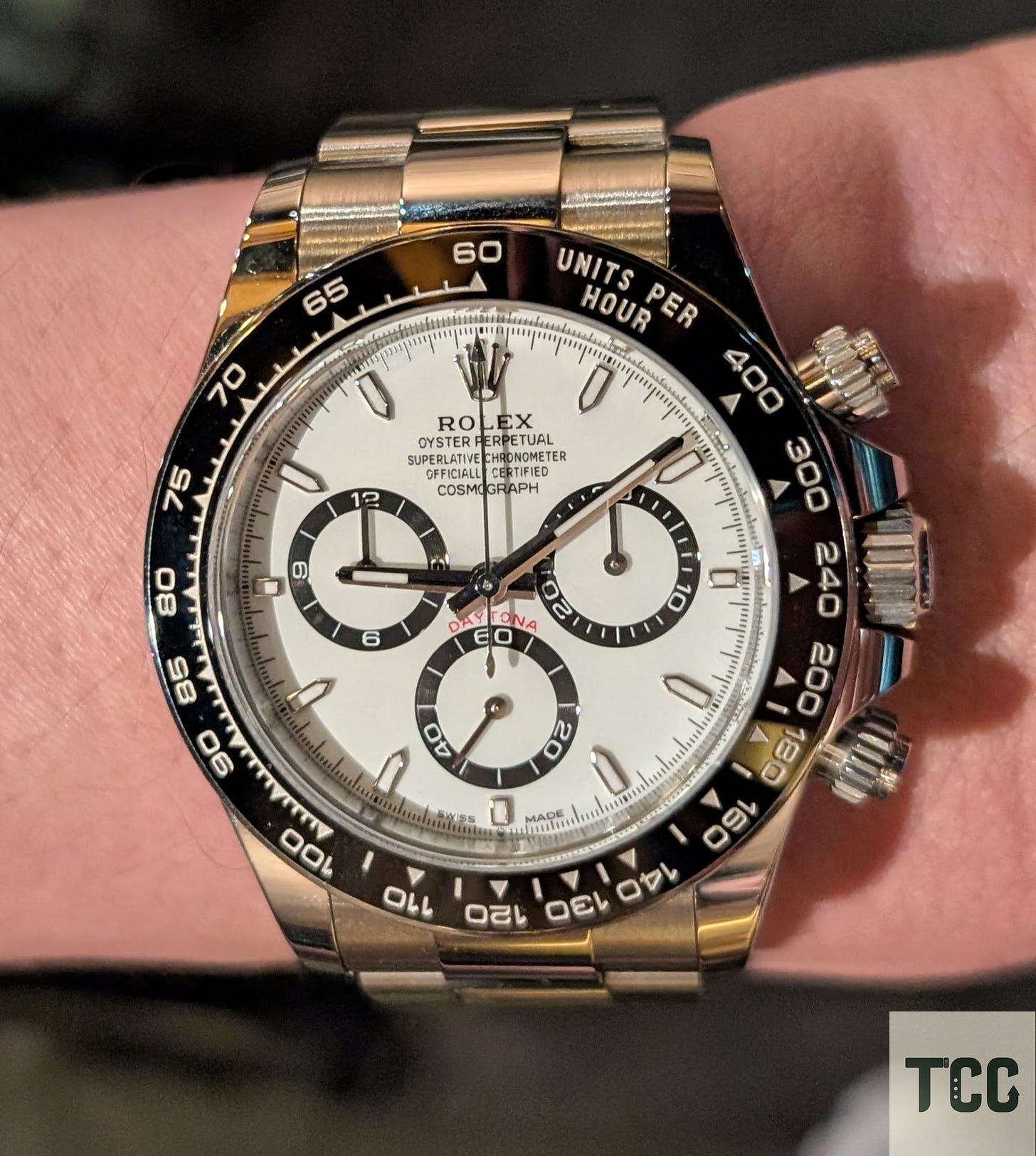 A close-up shot of a Rolex Daytona with a white dial, black ceramic bezel, and stainless steel bracelet. The subdials and red “Daytona” text stand out sharply, worn on the wrist of a fair-skinned hand.