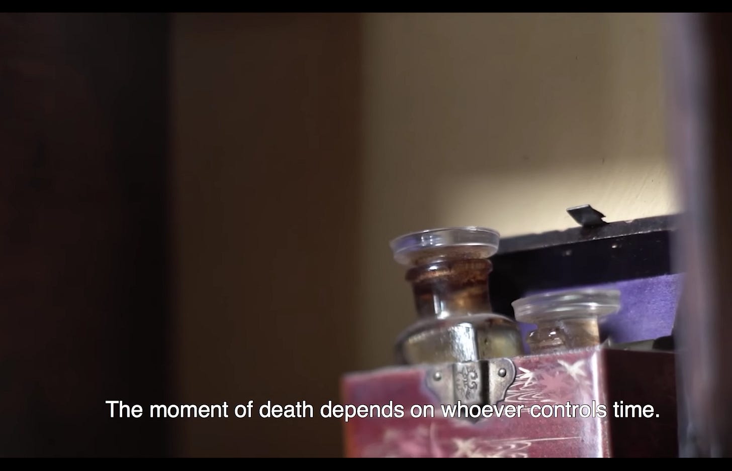 A film still with a closed caption that reads "The moment of death depends on whoever controls time." IN the background is a chest with two medicine bottles sticking out of the top. A film still with a closed caption that reads "The moment of death depends on whoever controls time." IN the background is a chest with two medicine bottles sticking out of the top.