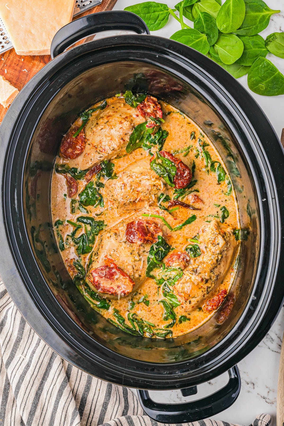 Chicken breasts cooked in a creamy sauce with spinach and sun-dried tomatoes in a slow cooker, with fresh spinach and parmesan cheese nearby.