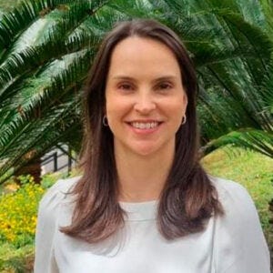 mulher de cabelos castanhos e compridos, usando uma blusa branca. Ela sorri para a câmera, em frente a um jardim com vegetação verde ao fundo mulher de cabelos castanhos e compridos, usando uma blusa branca. Ela sorri para a câmera, em frente a um jardim com vegetação verde ao fundo