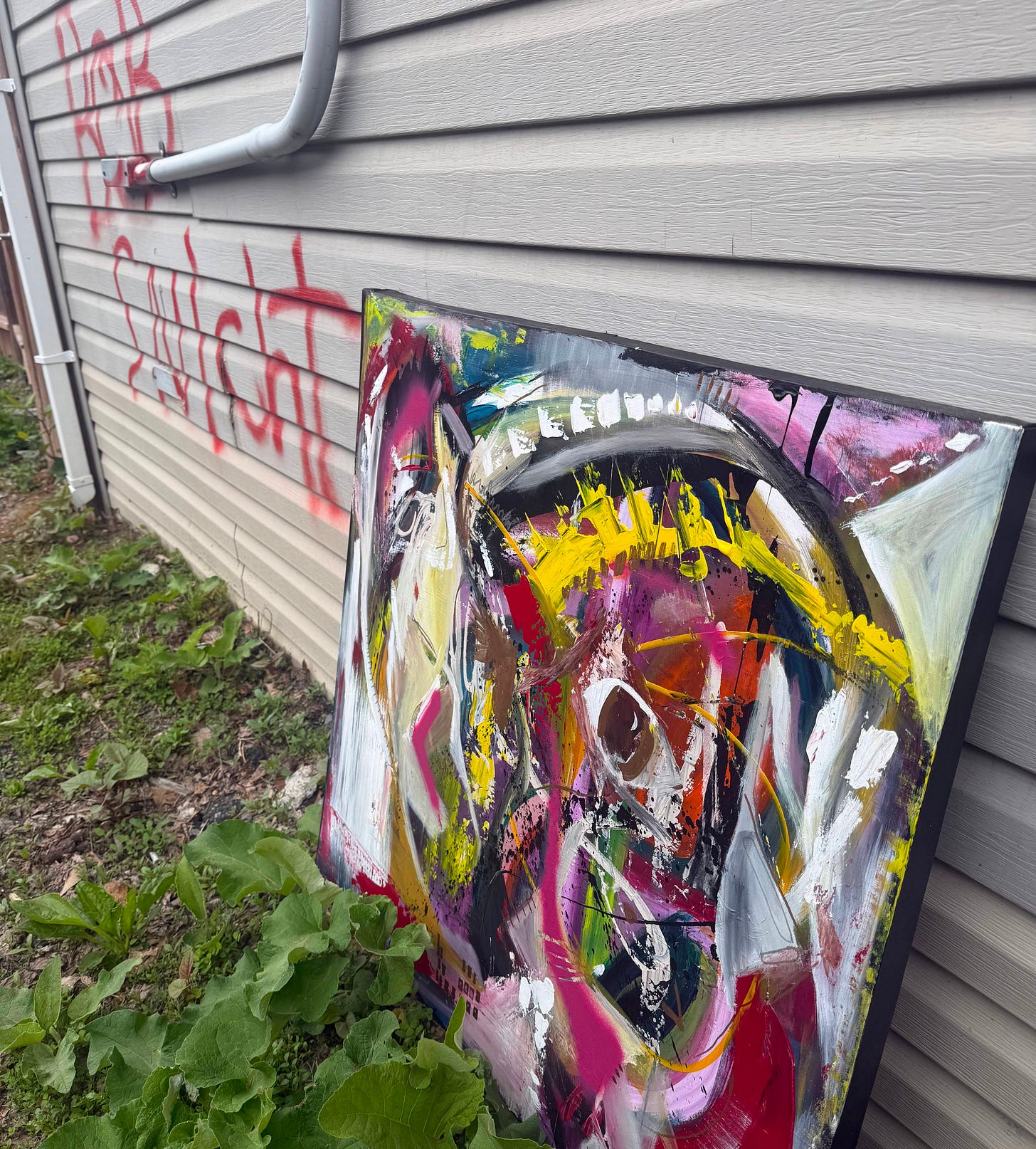 A large abstract painting leaning against the side of a building, partially obscured. Above it, the words BOB SNicht are spray-painted in red across the beige siding. Weeds grow at the base of the wall.