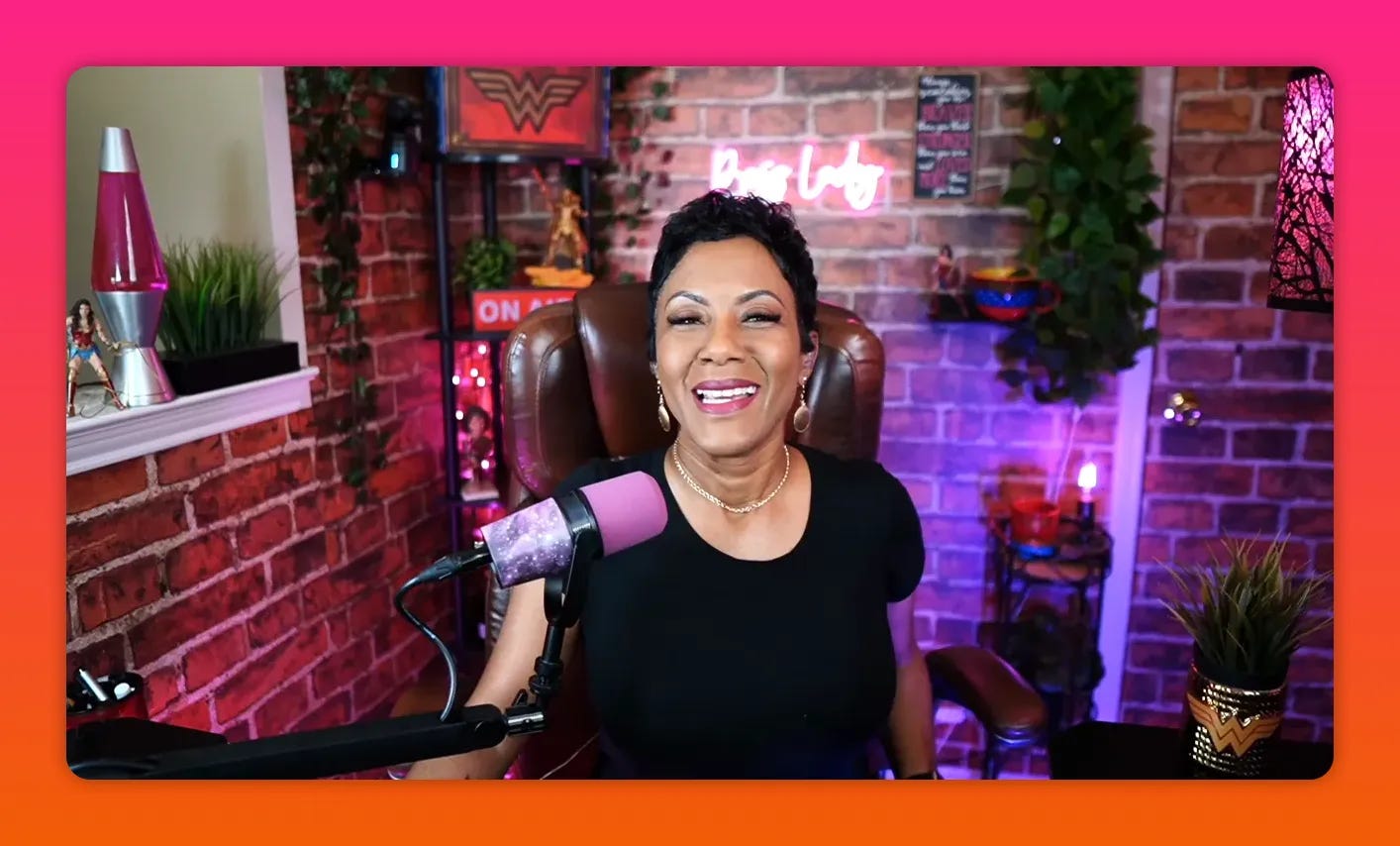Presenter smiling broadly in a well-lit brick-walled studio with a pink microphone and neon background sign.