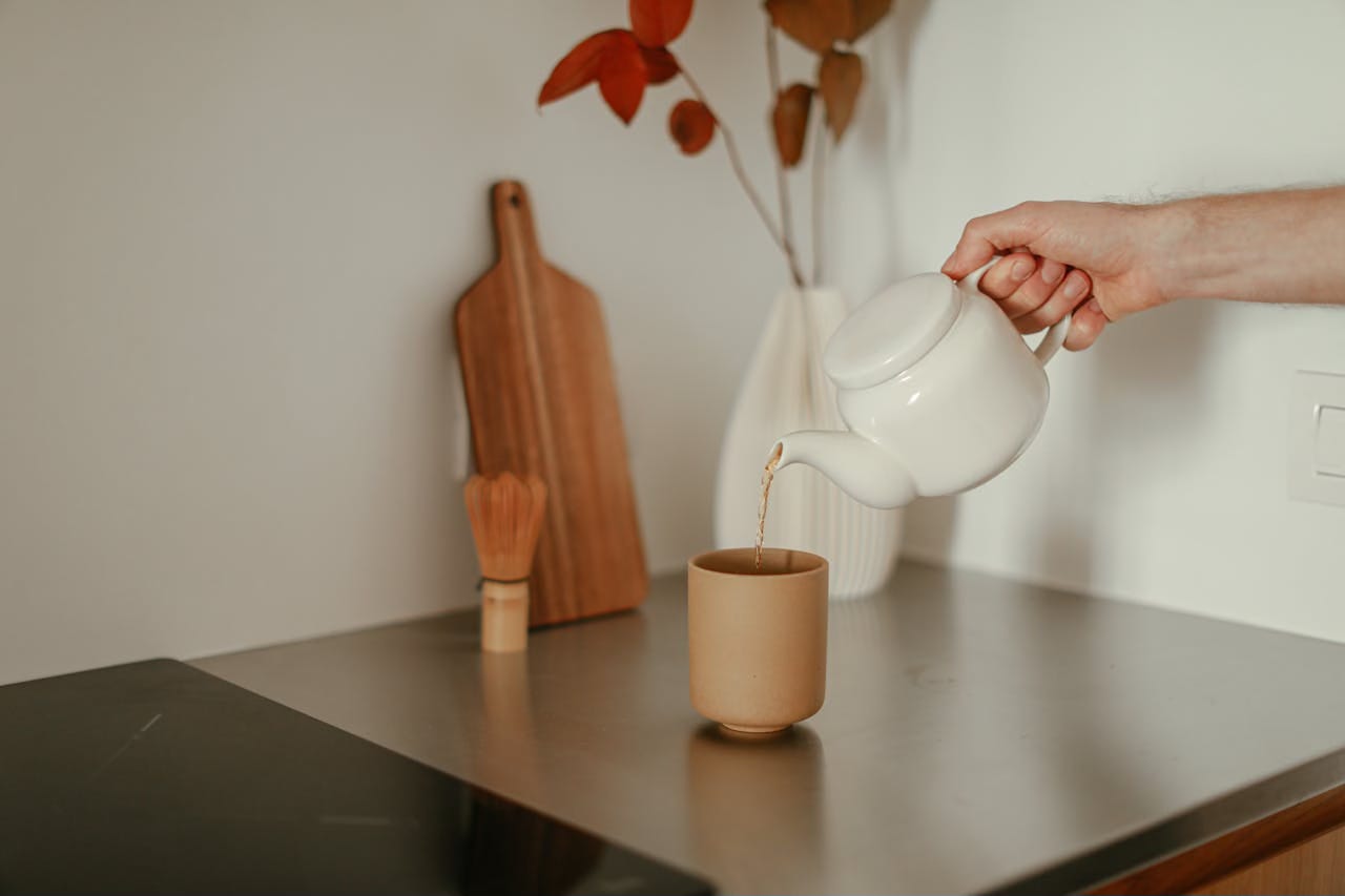 Arm pouring tea into a teacup on a table with a vase of red leaves. Arm pouring tea into a teacup on a table with a vase of red leaves.