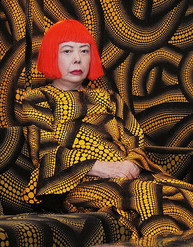The artist Yayoi Kusama with her signature orange wig, wearing a print that is repeated in the backdrop.