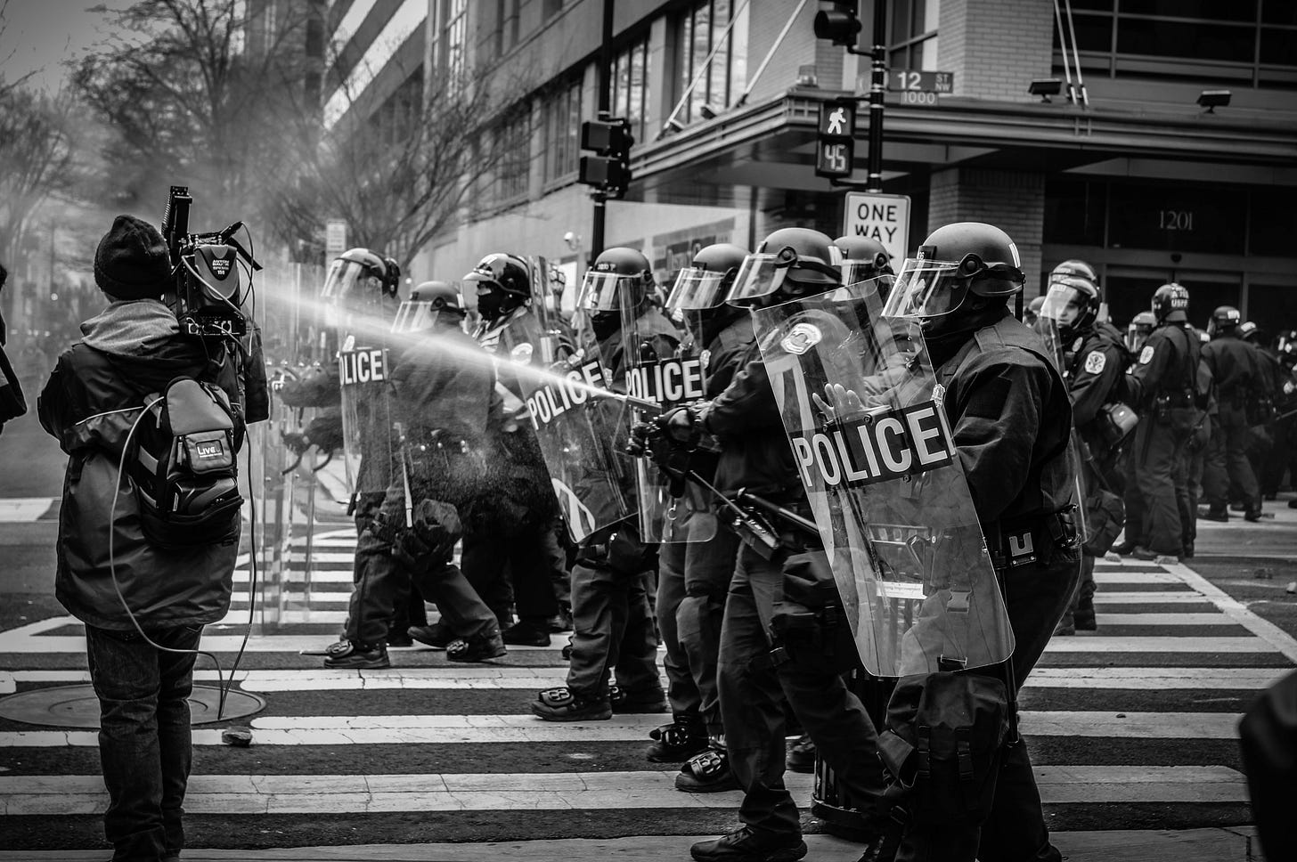 A grascale photo of police in riot gear spraying pepper spray at people out of frame with a camera operator right in front of them.