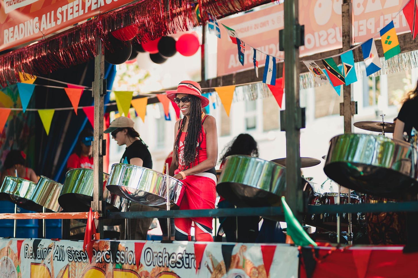 A vibrant steel band performance at Notting Hill Carnival, with musicians playing polished steel pans beneath colourful Caribbean flags and bunting, photographed by Rahimat Onize Shaibu.