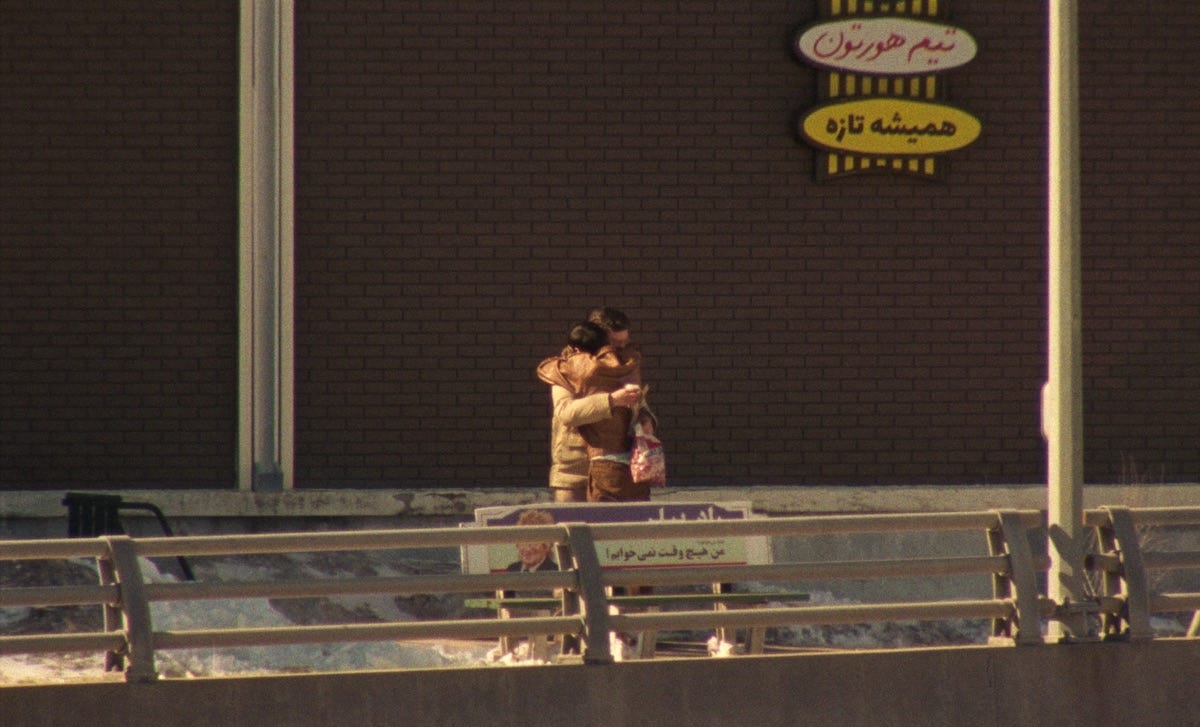 A film still depicts two people hugging behind a bench on a snowy walkway. Behind them is a brown brick wall that features a green, brown and white sign inscribed with Farsi using horizontal oval shapes.