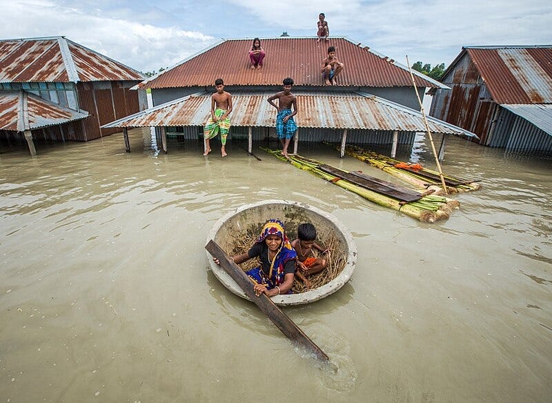 photo of a woman and young boy floating in a large round bowl-like wooden vessel. She uses a length of two-by-four as an oar. Behind them are three houses flooded nearly to the height of their rusty corrugated metal roofs, where several young men and children are sitting above the floodwaters. photo of a woman and young boy floating in a large round bowl-like wooden vessel. She uses a length of two-by-four as an oar. Behind them are three houses flooded nearly to the height of their rusty corrugated metal roofs, where several young men and children are sitting above the floodwaters.