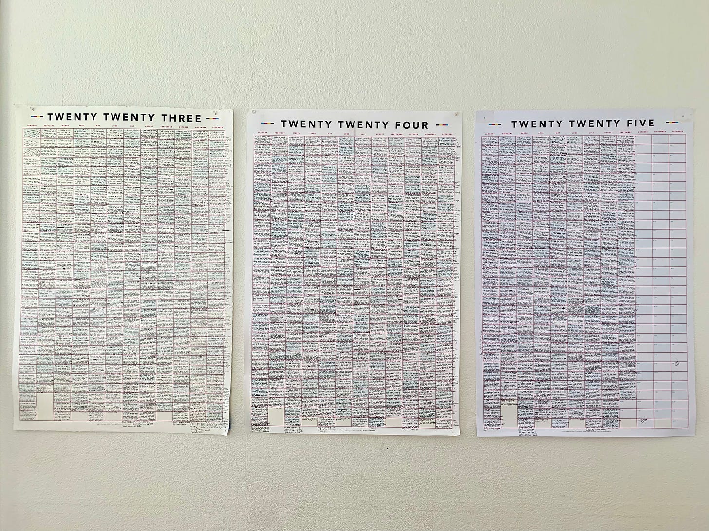 Three giant calendars pinned to a blank white wall. On the far left, the top of the calendar has the words “TWENTY TWENTY THREE,” in the center “TWENTY TWENTY FOUR,” and on the far right “TWENTY TWENTY FIVE.” Every inch of the calendars are filled with teenty tiny text except the last three months of 2025.