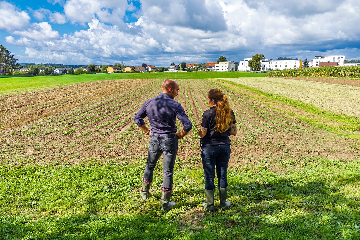 Zwei Studierende in Gummistiefeln stehen auf dem Oberacker in Zollikofen und blicken über frisch bestellte Reihen eines Versuchsfeldes. Im Hintergrund sind weitere Felder, Maiskulturen sowie Wohngebäude am Ortsrand zu sehen, darüber ein Himmel mit imposanten Wolkenformationen.
