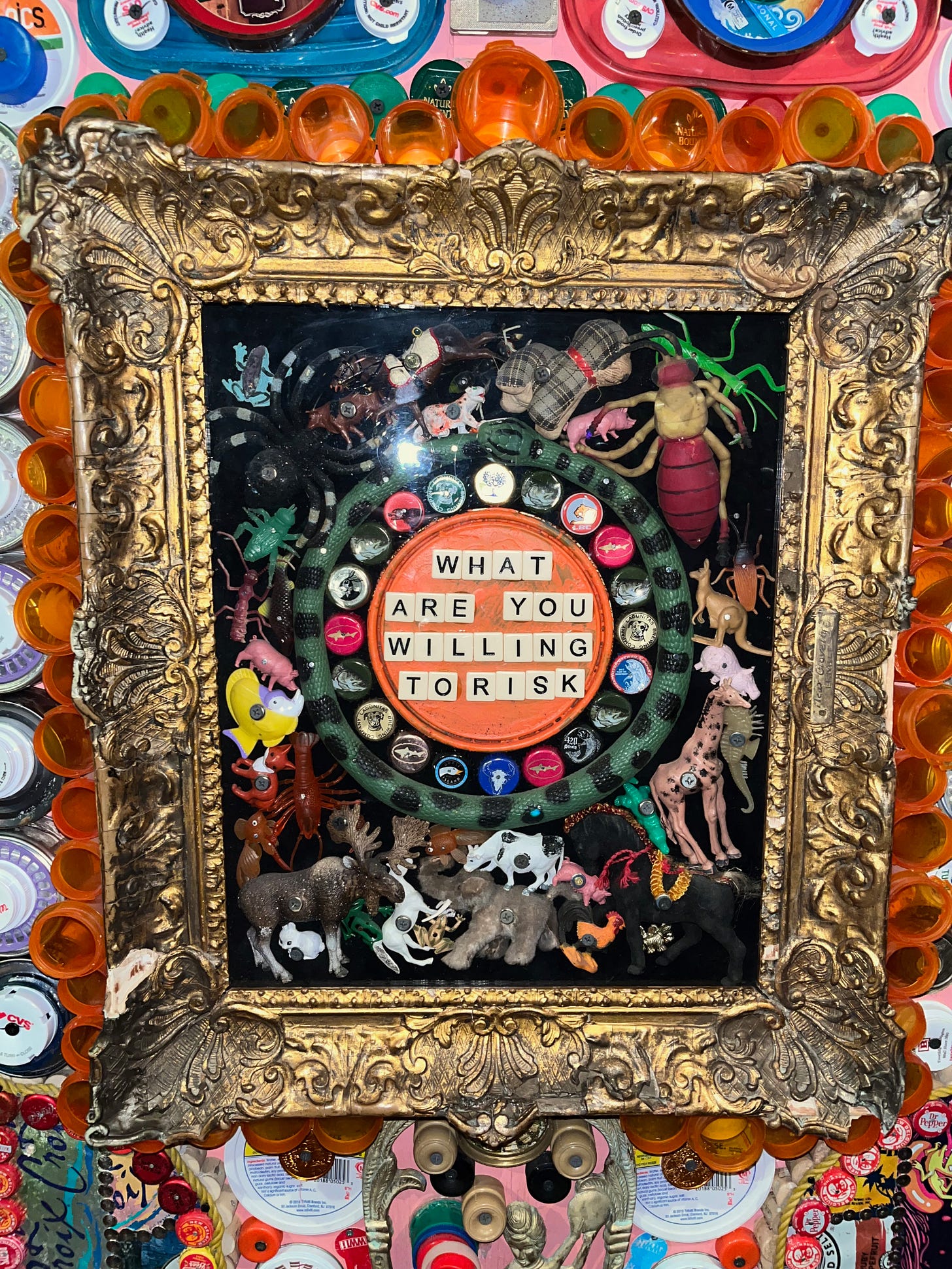 Art piece from Meow Wolf, Santa Fe. Circle with Scrabble letters spelling out "what are you willing to risk" surrounded by figurines of plastic animals in a gold frame on a decorated wall.