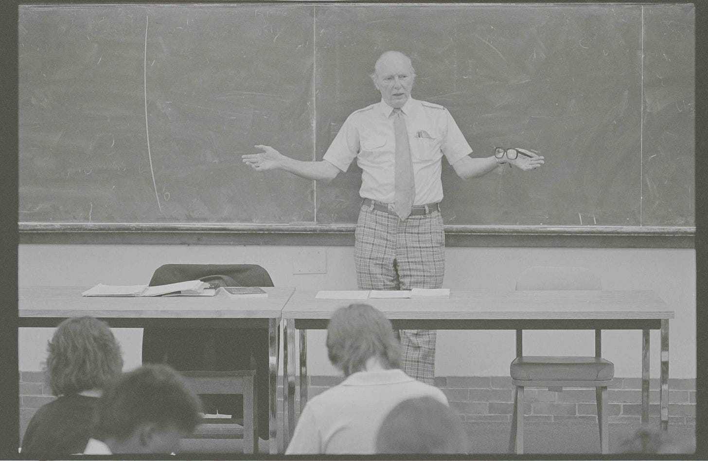 Black and white image of University of Rochester political scientist William Riker, who codeveloped the Riker-Ordeshook theory of voter turnout behavior, gestures at the front of a classroom.