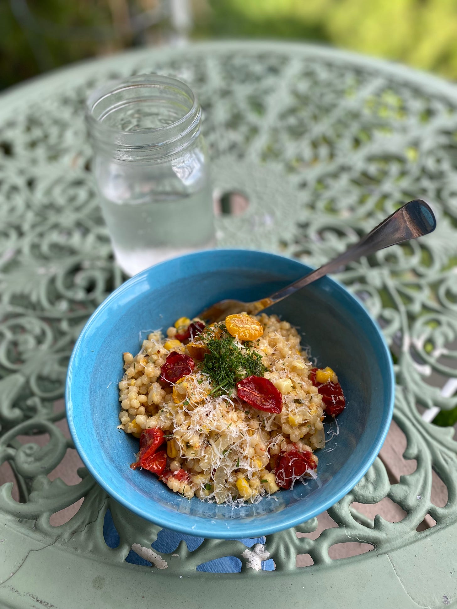 A blue bowl of the cousous described above, with multicolour roasted tomatoes, bits of corn, and a dusting of parmesan and mixed herbs. We're at the outdoor cafe table again, a sweating jar of water sitting behind the bowl.