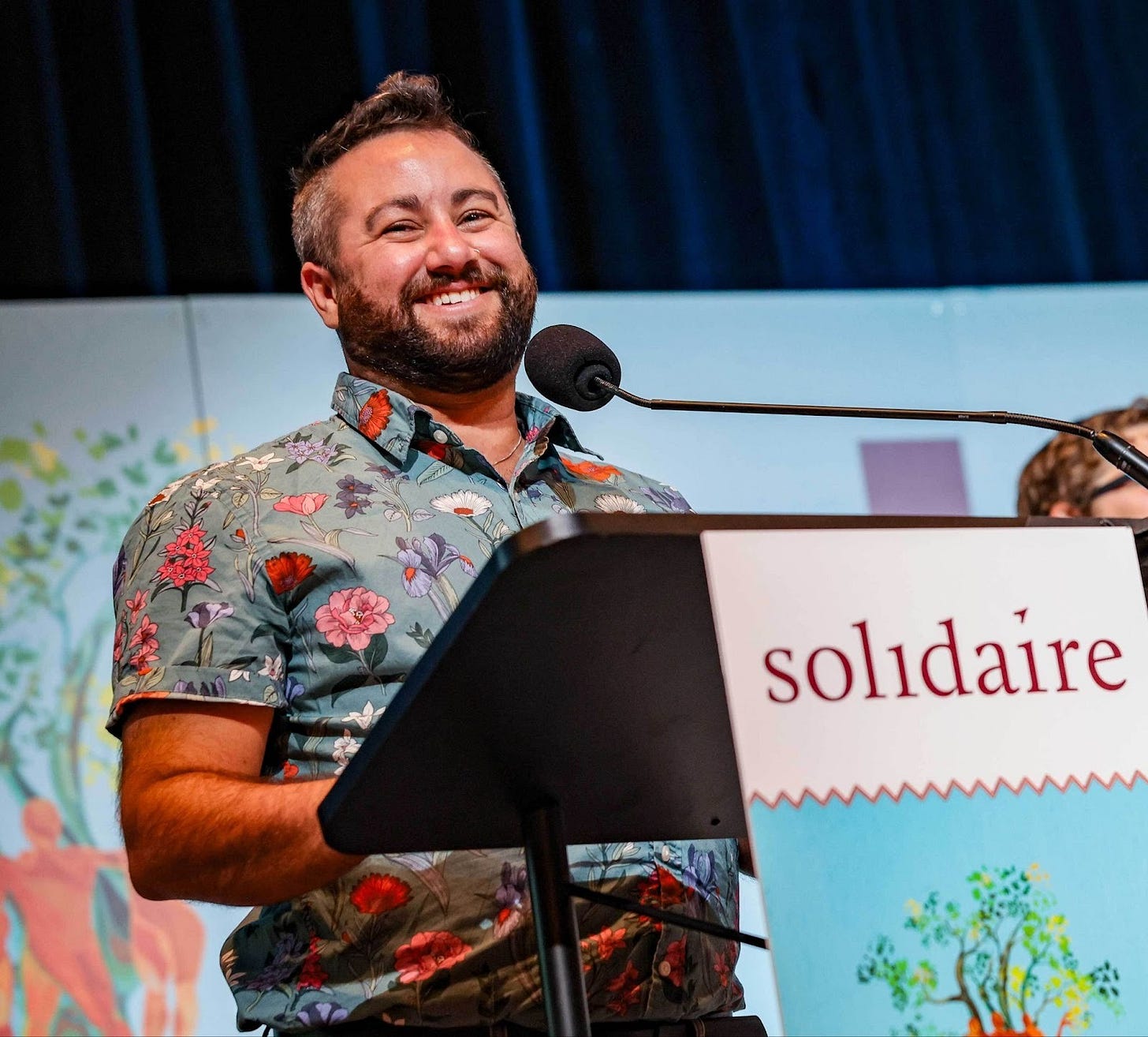 A photo of Isaac Lev, standing behind a lectern, wearing a floral print button up shirt, smiling, with olive skin, and short black and grey hair and a black/brown beard.