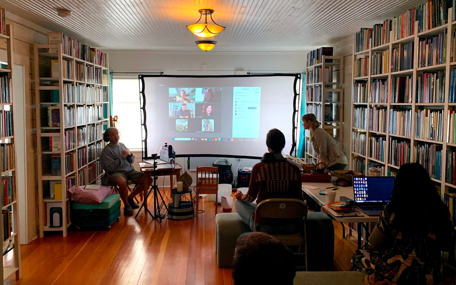 People learning in the main reading room at CAELi surrounded by astrology books, and facing a screen with zoom attendees beaming in to listen to the instructor, Cameron Cassidy.
