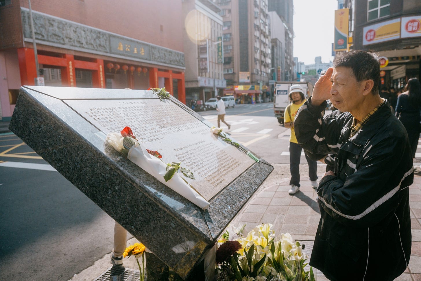 An elderly Asian man saluting a large metal and marble plaque on the side of a street. White flowers are seen at the base of the monument, with more flowers on the plaque itself. (Image contributed by Philip So)