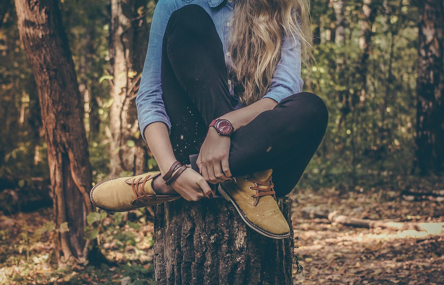 A cropped woman with long brown hair and denim shirt sits cross-legged atop a large tree stump. A cropped woman with long brown hair and denim shirt sits cross-legged atop a large tree stump.