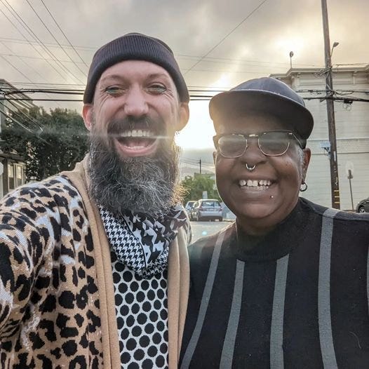 Jason and Crystal standing side-by-side with the sun shining through the fog behind them. Both are smiling wide and joyfully.