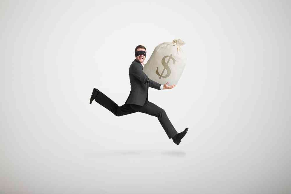 A man in a mask and black clothing leaps through the air while holding a huge bag of money.
