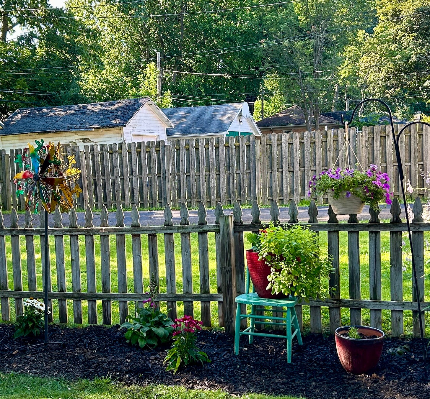 Old wooden picket fence with a flower garden in front. Garden contains spearmint, Gerber daisies, white and yellow daisies, foxglove, cone flowers, rosemary, and petunias. There's a sweet little light green chair that I found on the curb one day supporting a pot. There's also a fun colorful metal pinwheel.