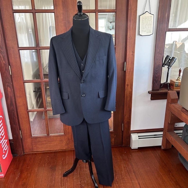 May include: A navy blue pinstripe suit, including a jacket, vest, and trousers, displayed on a black mannequin. The suit has a classic three-button closure on the jacket and a matching vest. The trousers are tailored with a straight leg. The suit is displayed on a wooden floor.
