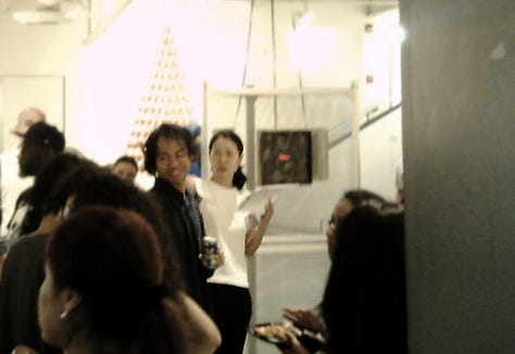 A slightly (but artfully) blurry photo that shows several people looking on at Souya Handa and NJCU art department chair in the brightly lit Visual Arts Building Gallery, where the red LED light of one of Handa's pieces can be seen in the background.
