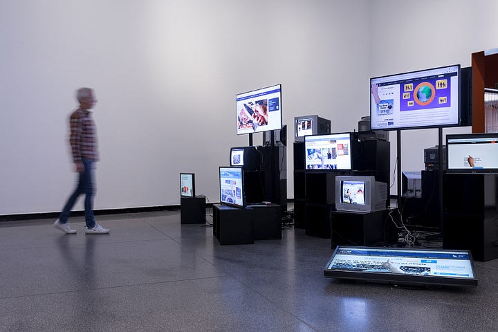 Undici monitor di computer, la maggior parte posizionati su alti piedistalli neri in un'ampia stanza con finestre dal pavimento al soffitto, espongono pagine web, video e contenuti digitali.