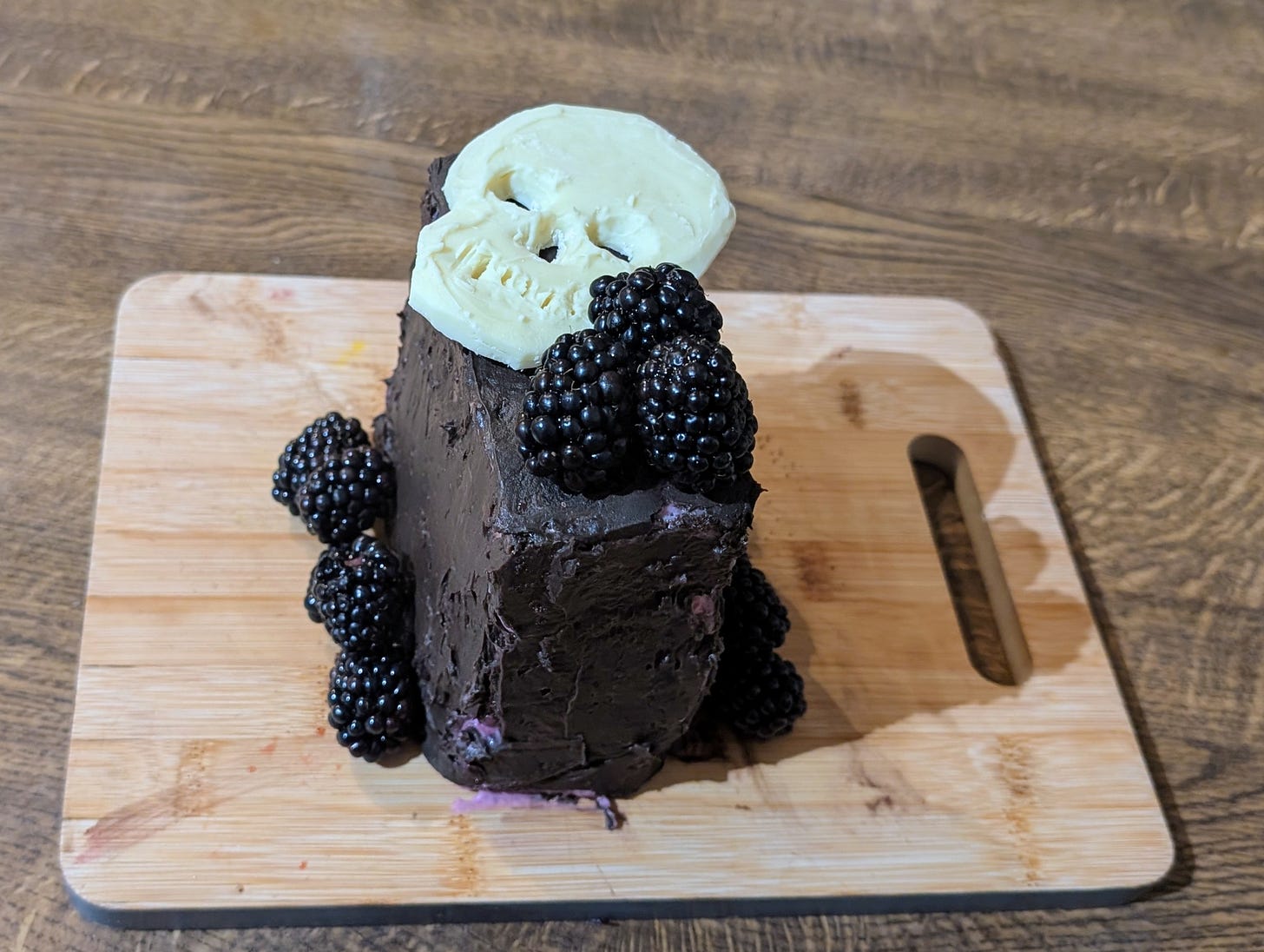 A small rectangular cake sits on a wooden board, coated in glossy dark chocolate frosting. Fresh blackberries ring the base and cluster on top, their deep purple almost merging with the icing. Centered on the cake is a white chocolate skull, its smooth surface stark against the dark, moody palette.